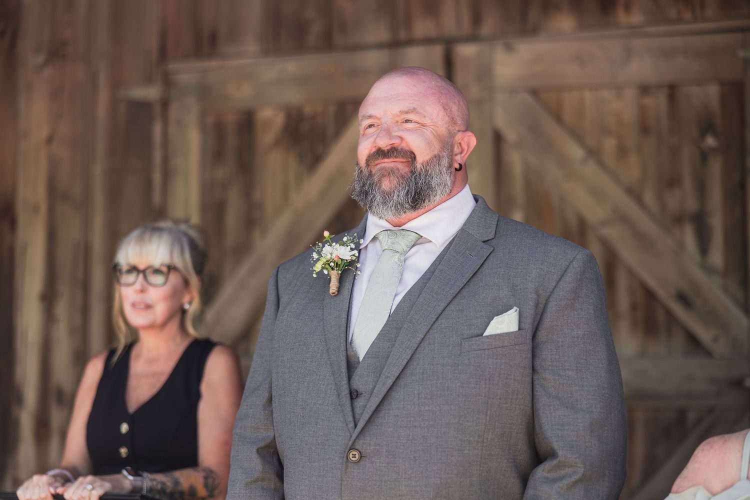 Bald groom with beard in gray suit and boutonniere smiling at wedding ceremony in rustic barn venue.