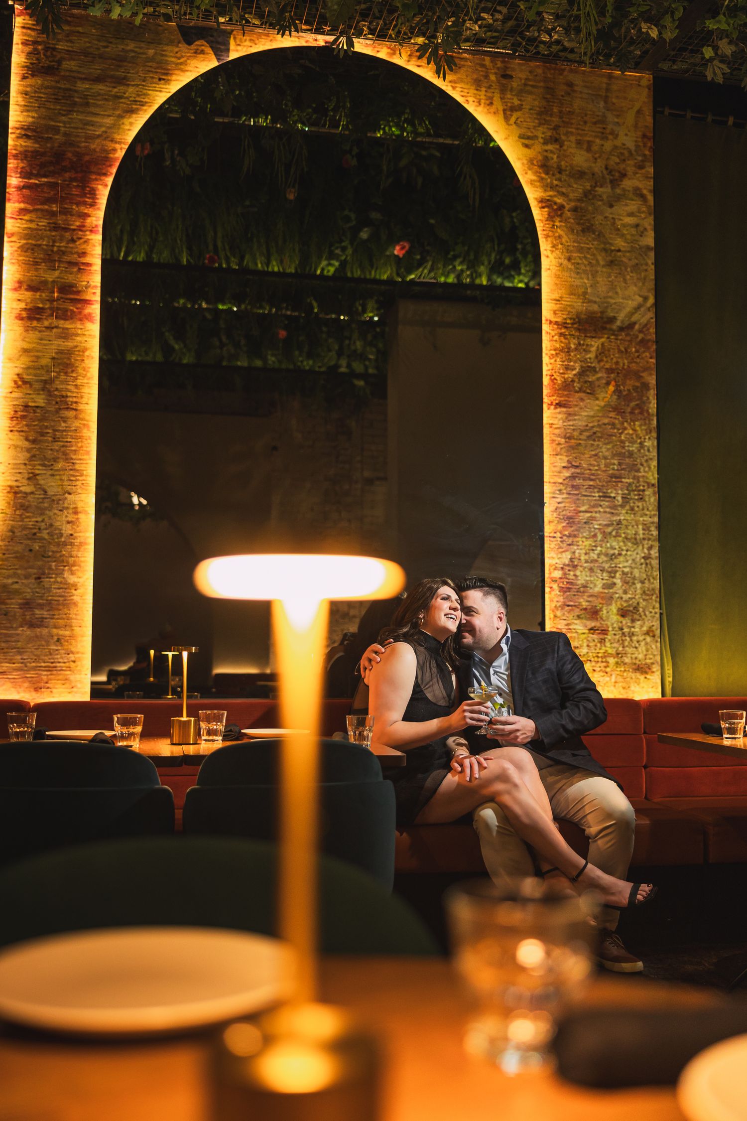 Couple sharing a romantic moment on a velvet sofa in a warmly lit restaurant with an arched brick backdrop.
