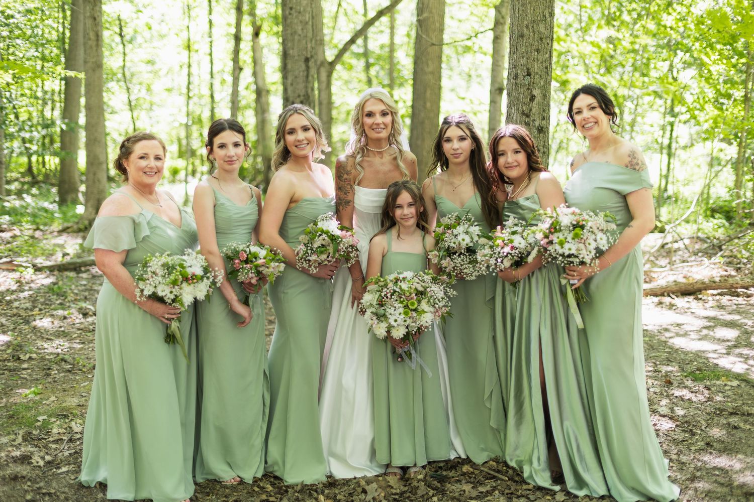 Bride poses with six bridesmaids in sage green gowns holding white bouquets in a forest setting.