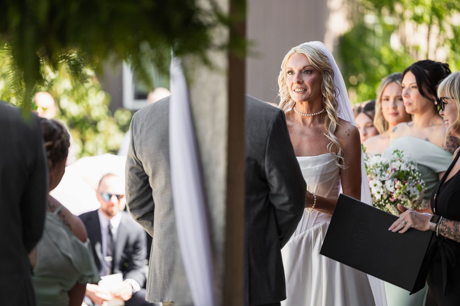 Elegant blonde bride in white gown smiles during outdoor wedding ceremony.