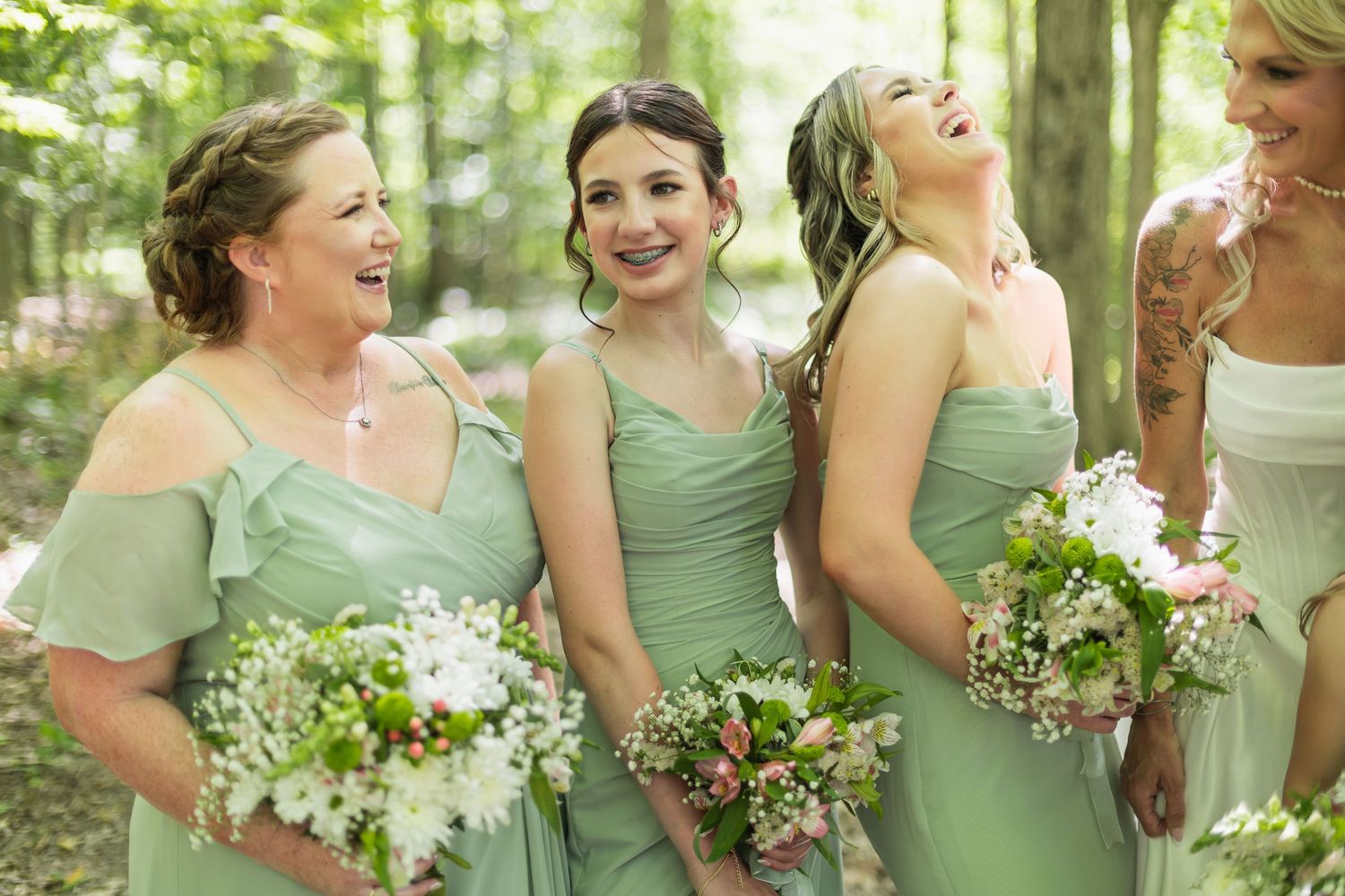 Four bridesmaids in sage green dresses holding white floral bouquets laughing together outdoors in a wooded setting.
