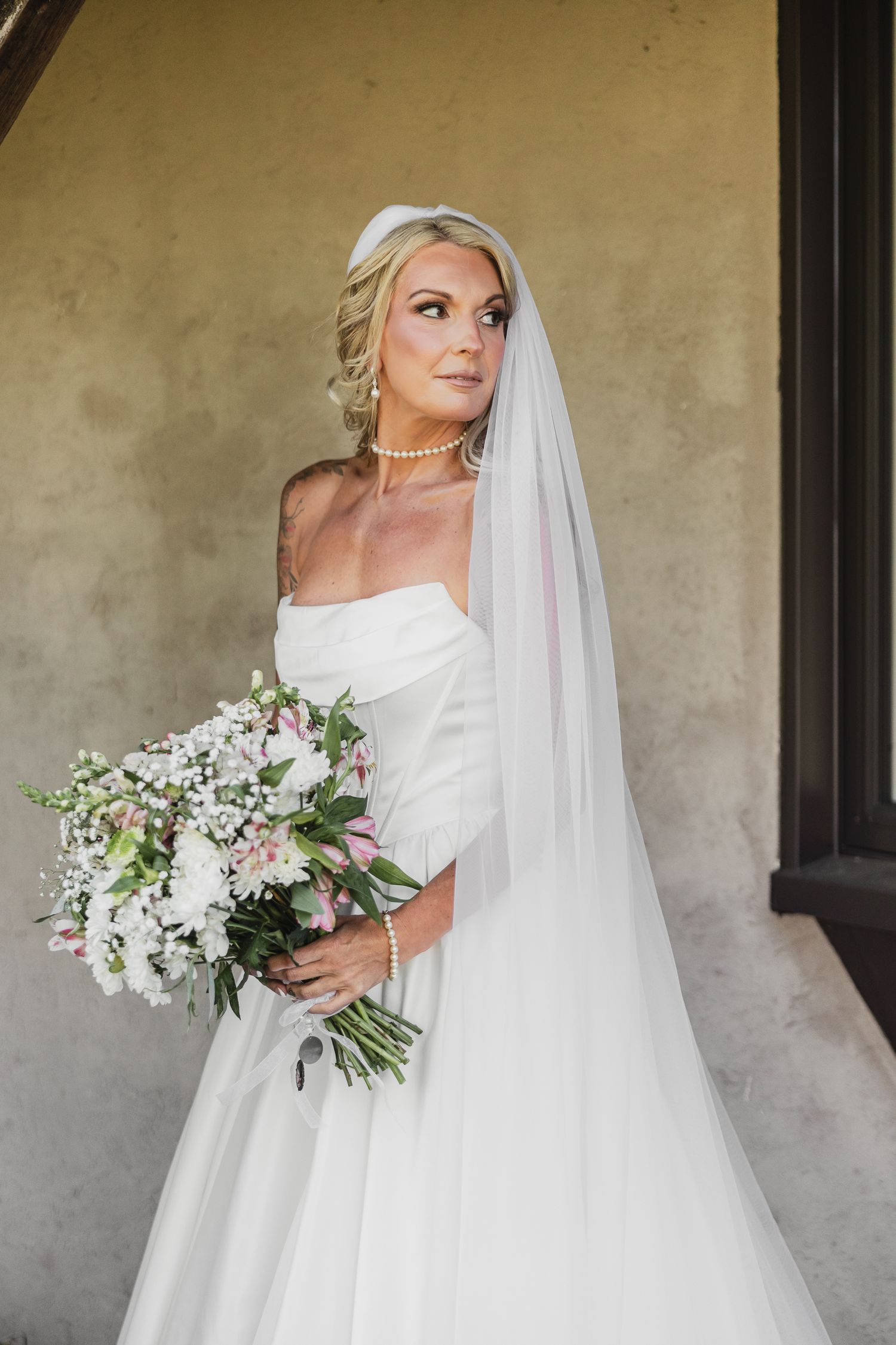 Elegant blonde bride in off-shoulder gown with veil and pearls holds pink and white bouquet.