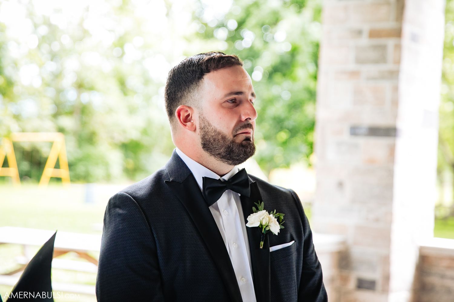 Groomsmen in black tuxedos stand together on a porch with brick columns and greenery in background.