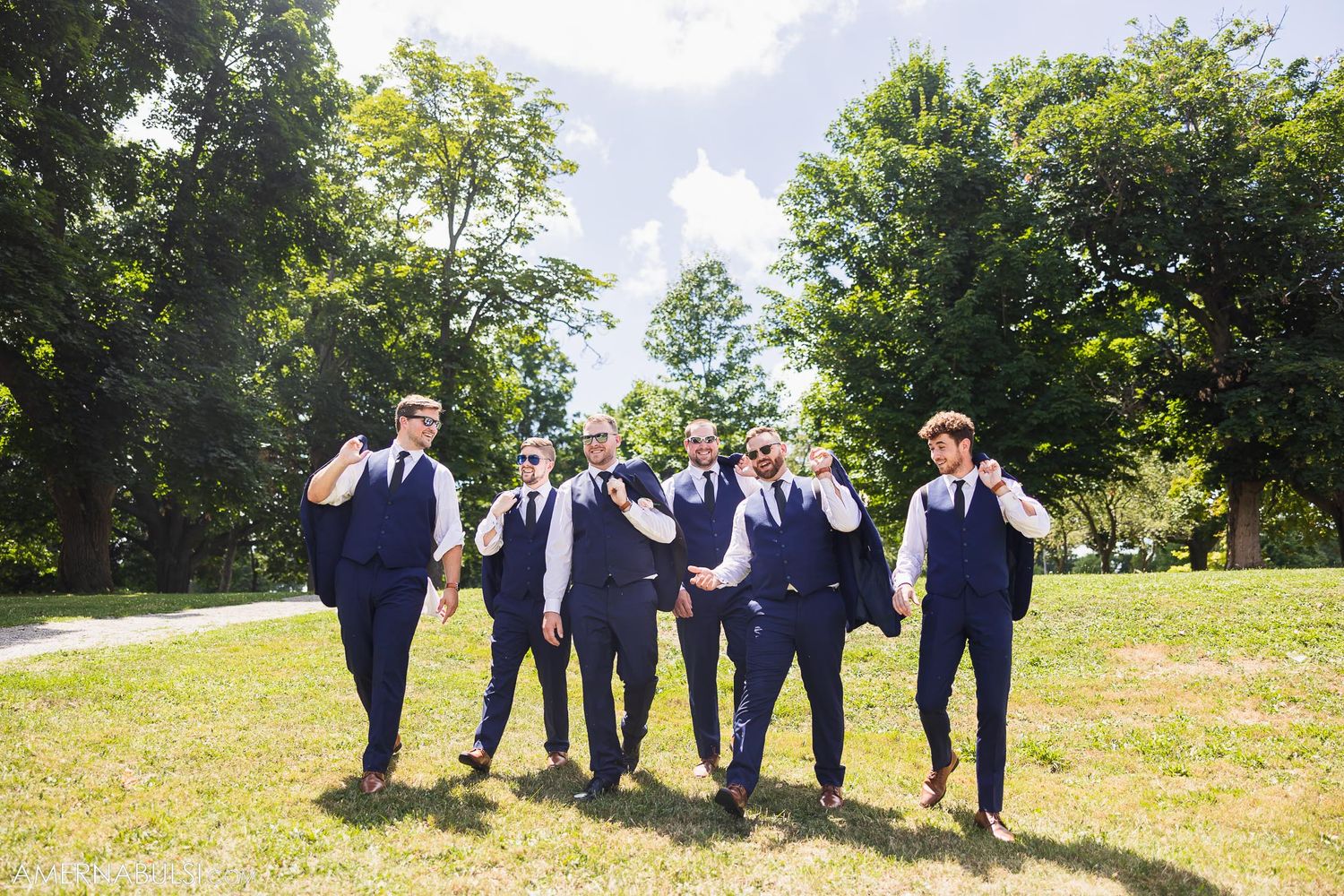 Groomsmen in matching navy vests walking casually across sunny lawn.