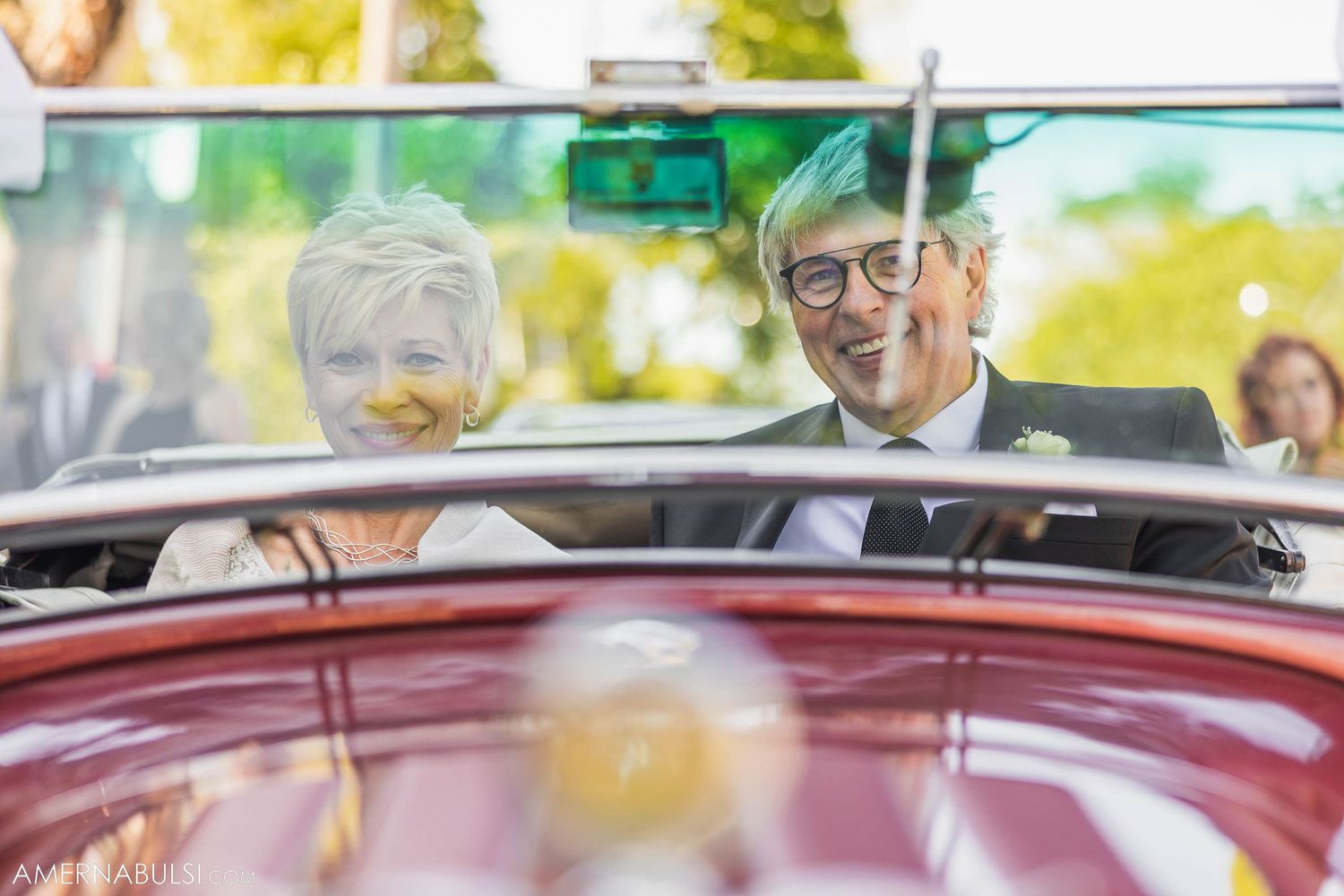 A couple riding in a vintage burgundy convertible car on a sunny day.