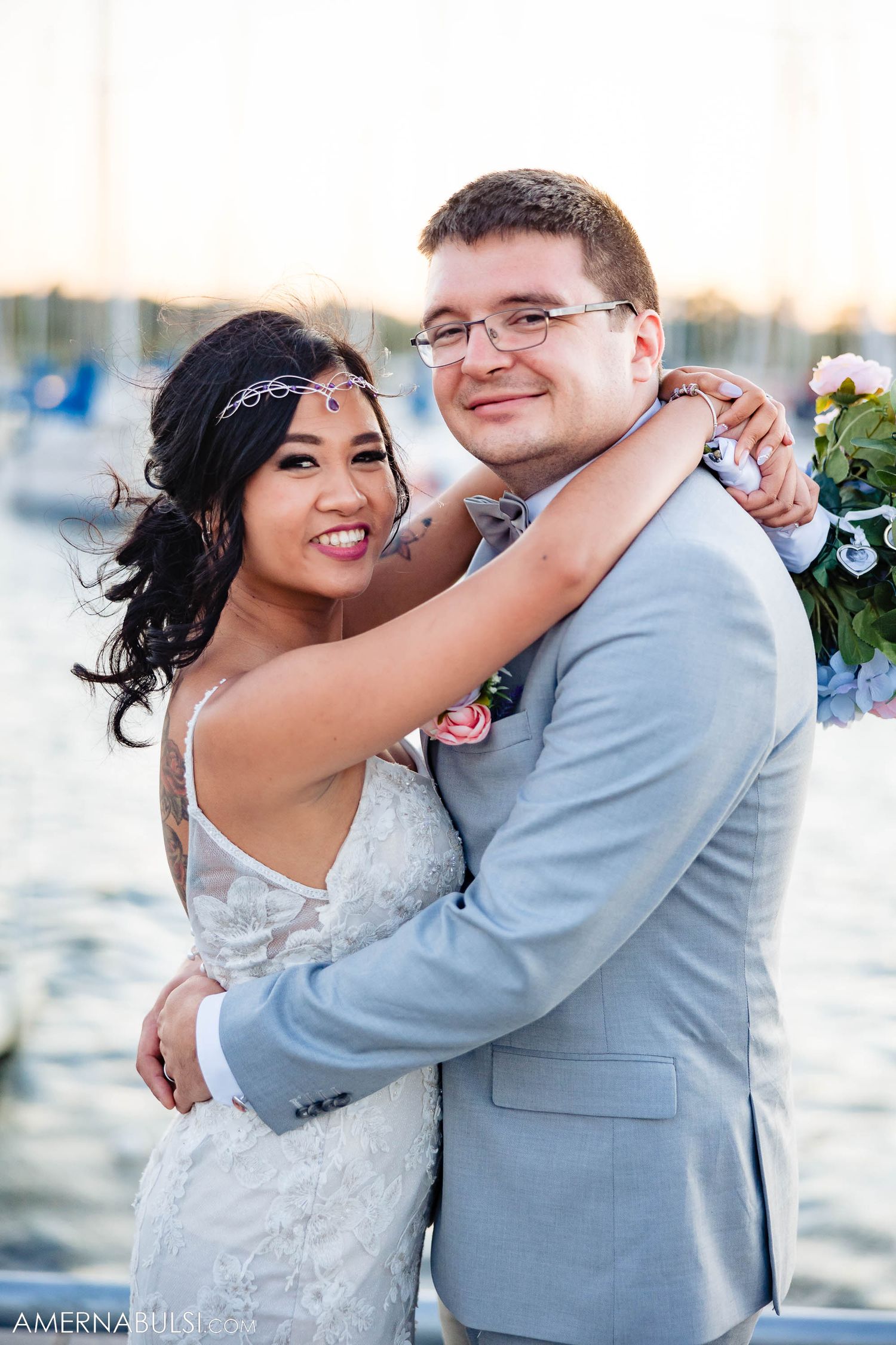 Newlyweds in light blue suit and white dress embrace by waterfront marina.
