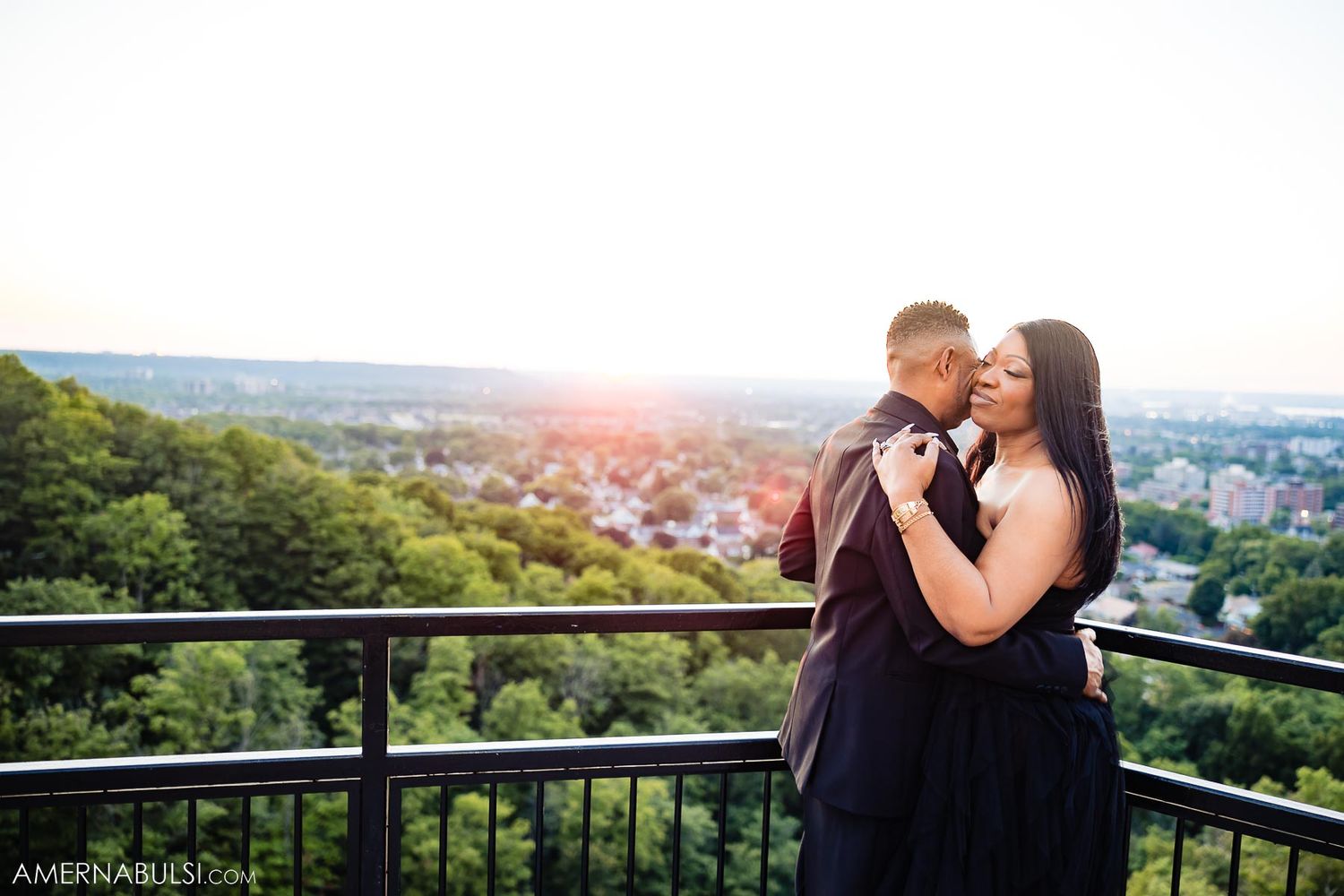 Couple embrace on observation deck overlooking city during sunset.
