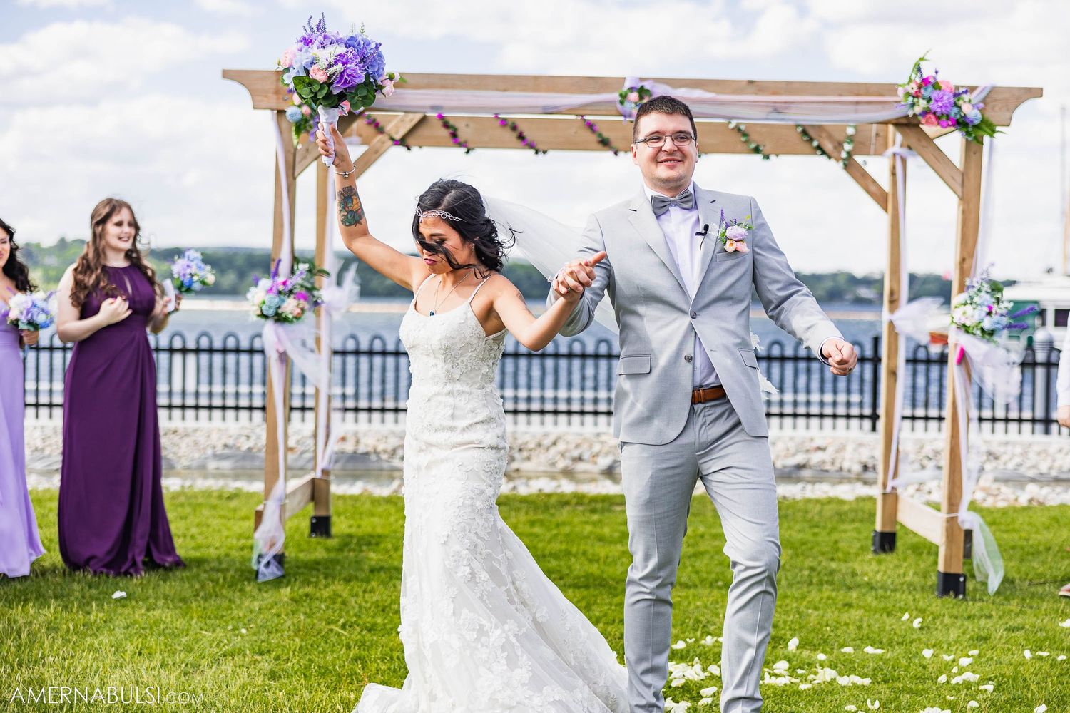 Joyful celebration under wooden wedding arch with purple floral decorations by the water.