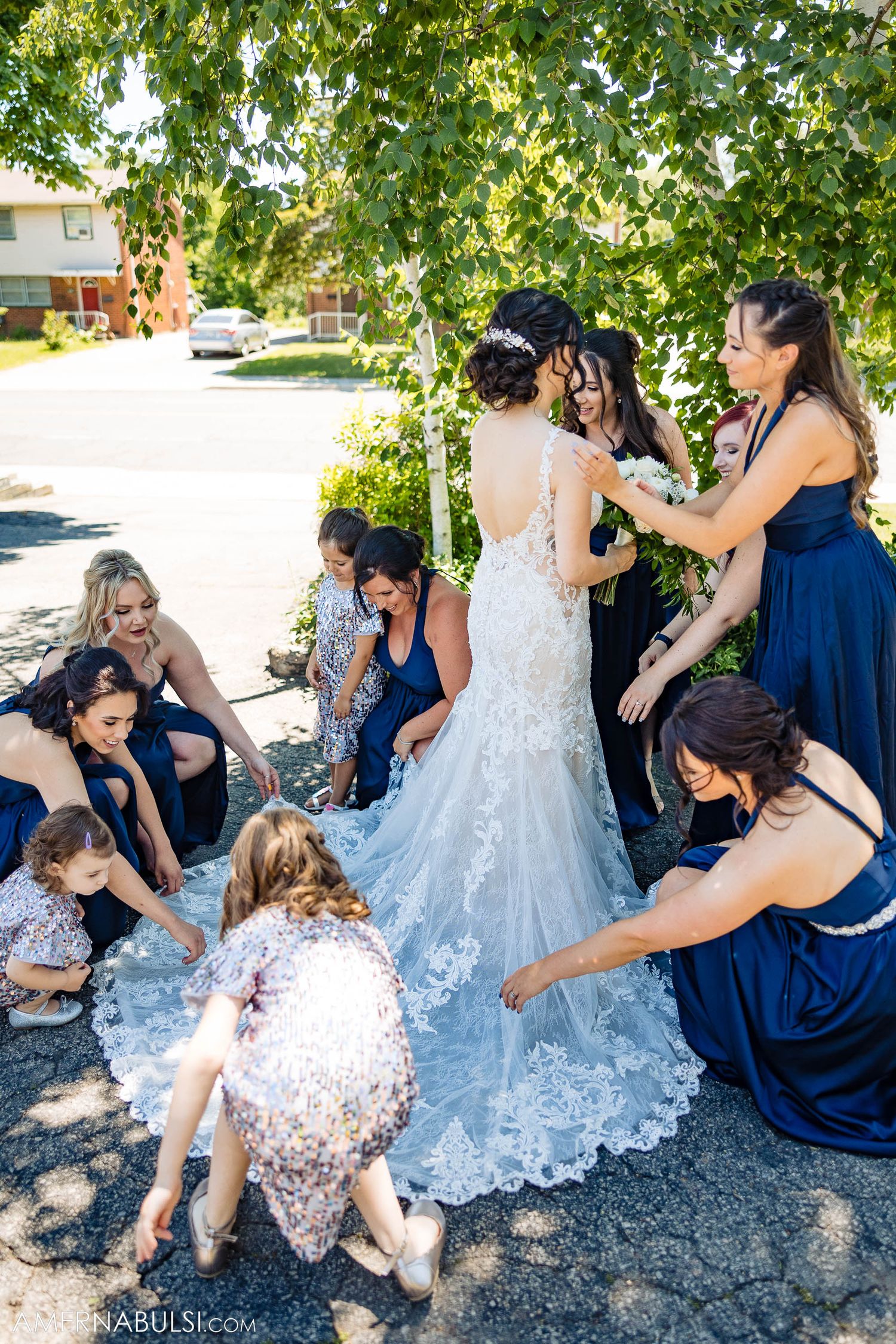 Bridesmaids in navy dresses helping arrange wedding gown train outdoors.