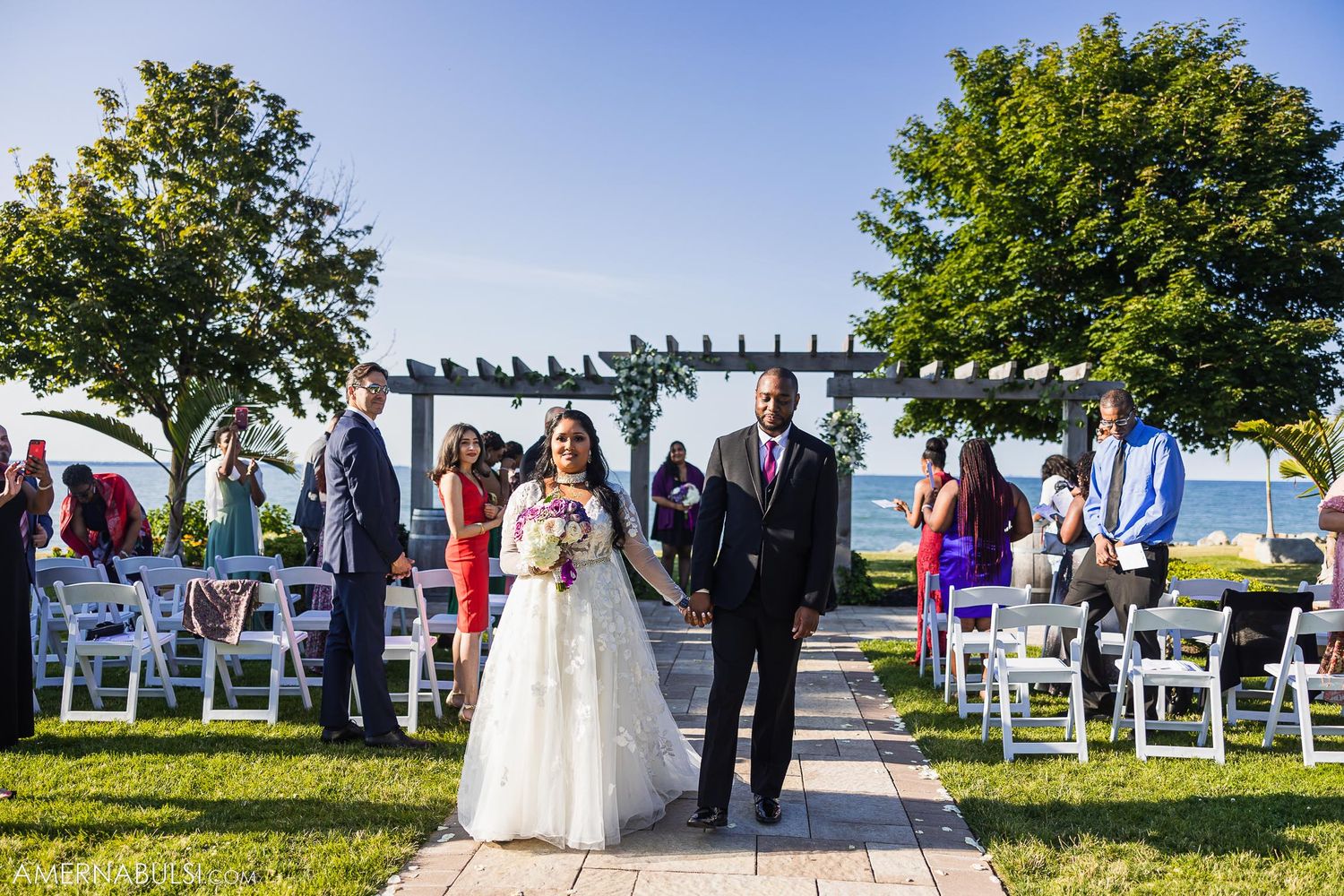 A couple walks down the aisle during an outdoor wedding ceremony with white chairs and a waterfront view.