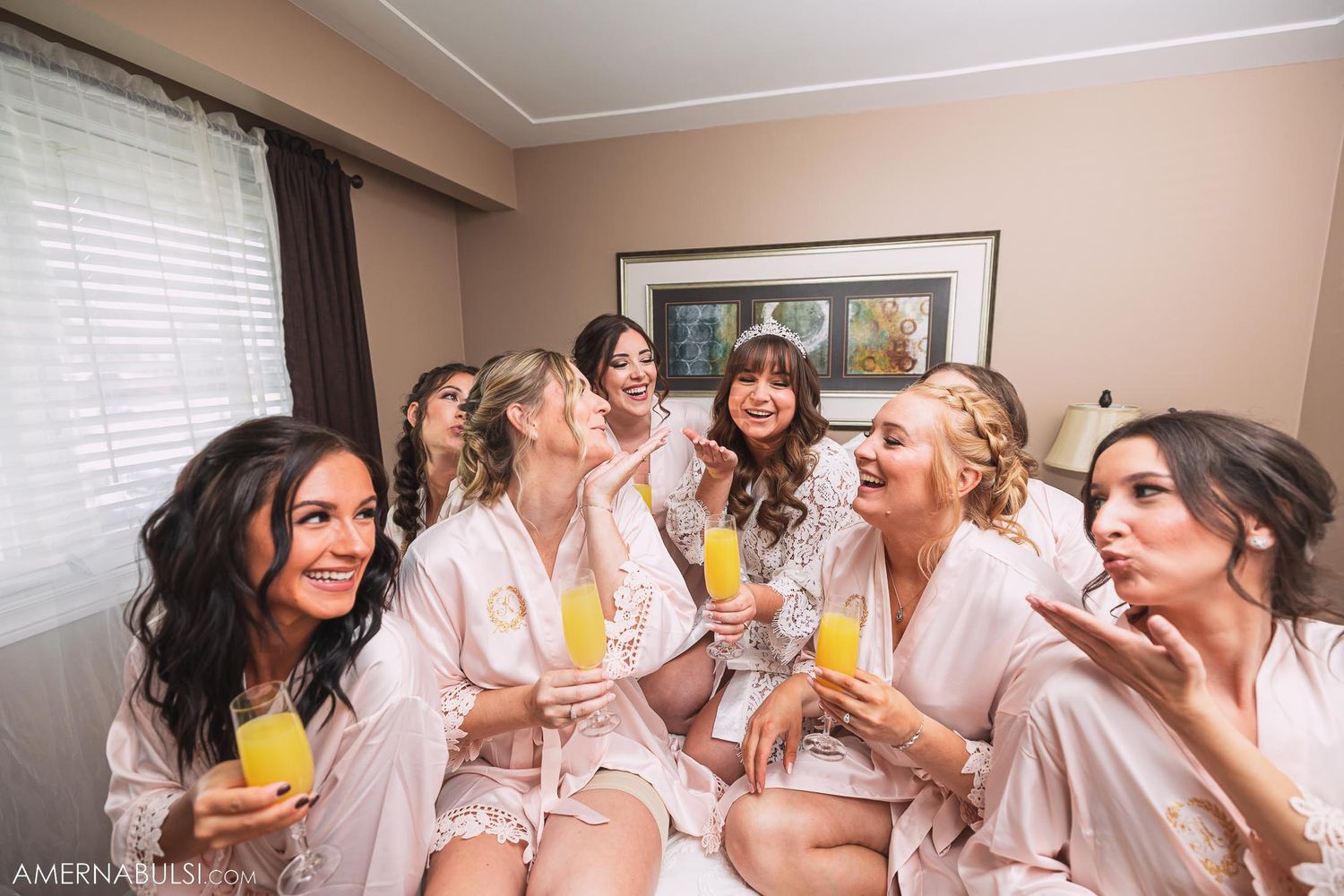 Bridesmaids in silk robes laugh together while enjoying drinks during pre-wedding preparations.