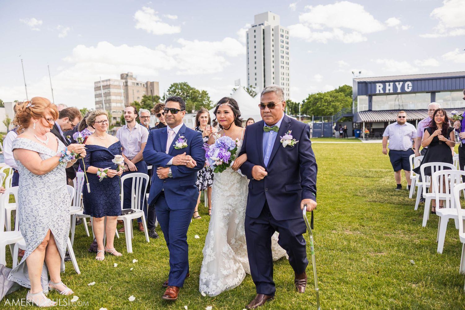 Wedding party walks across lawn with high-rise buildings and guests in background.