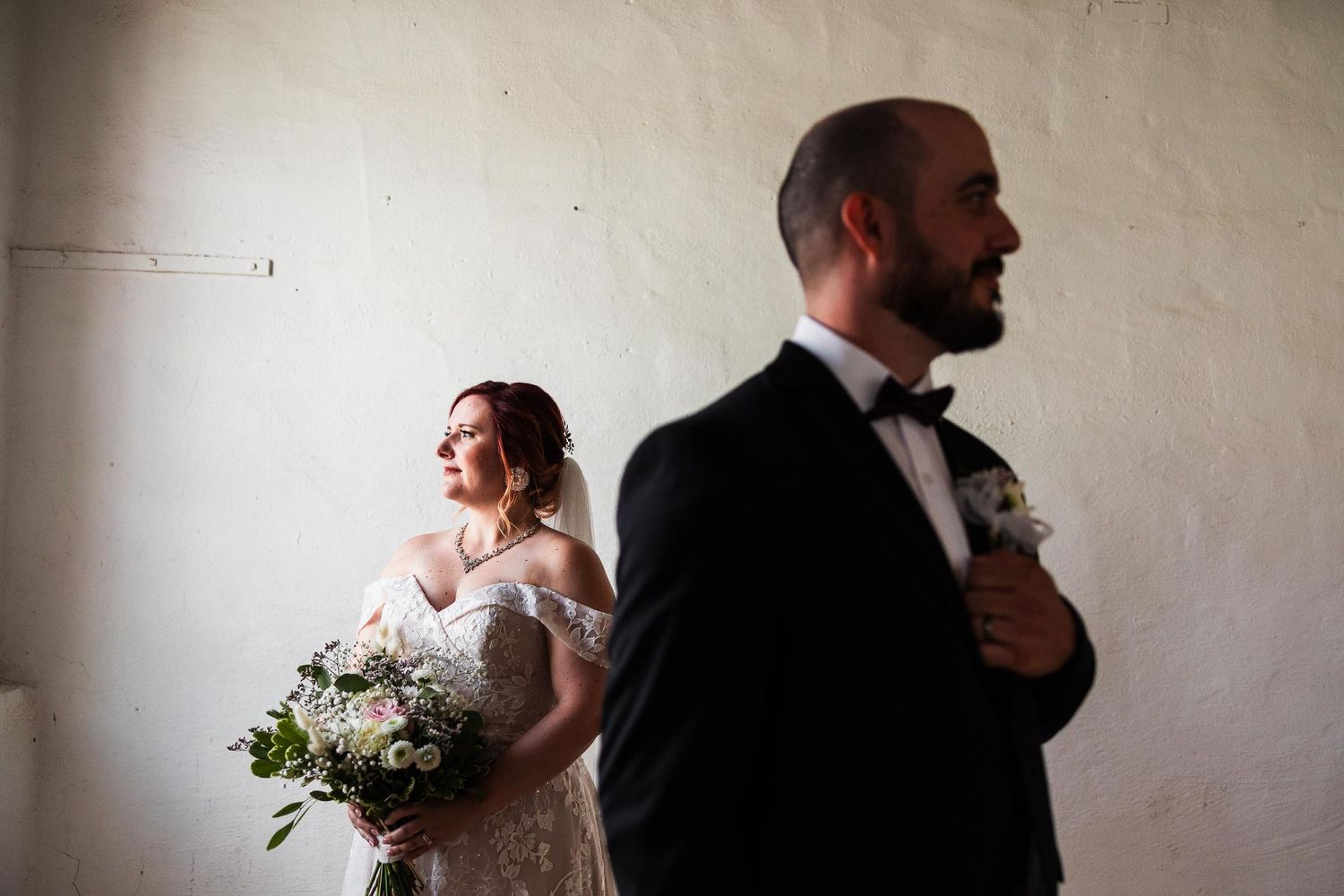A wedding couple stands apart in a minimalist room with the bride holding a white bouquet.
