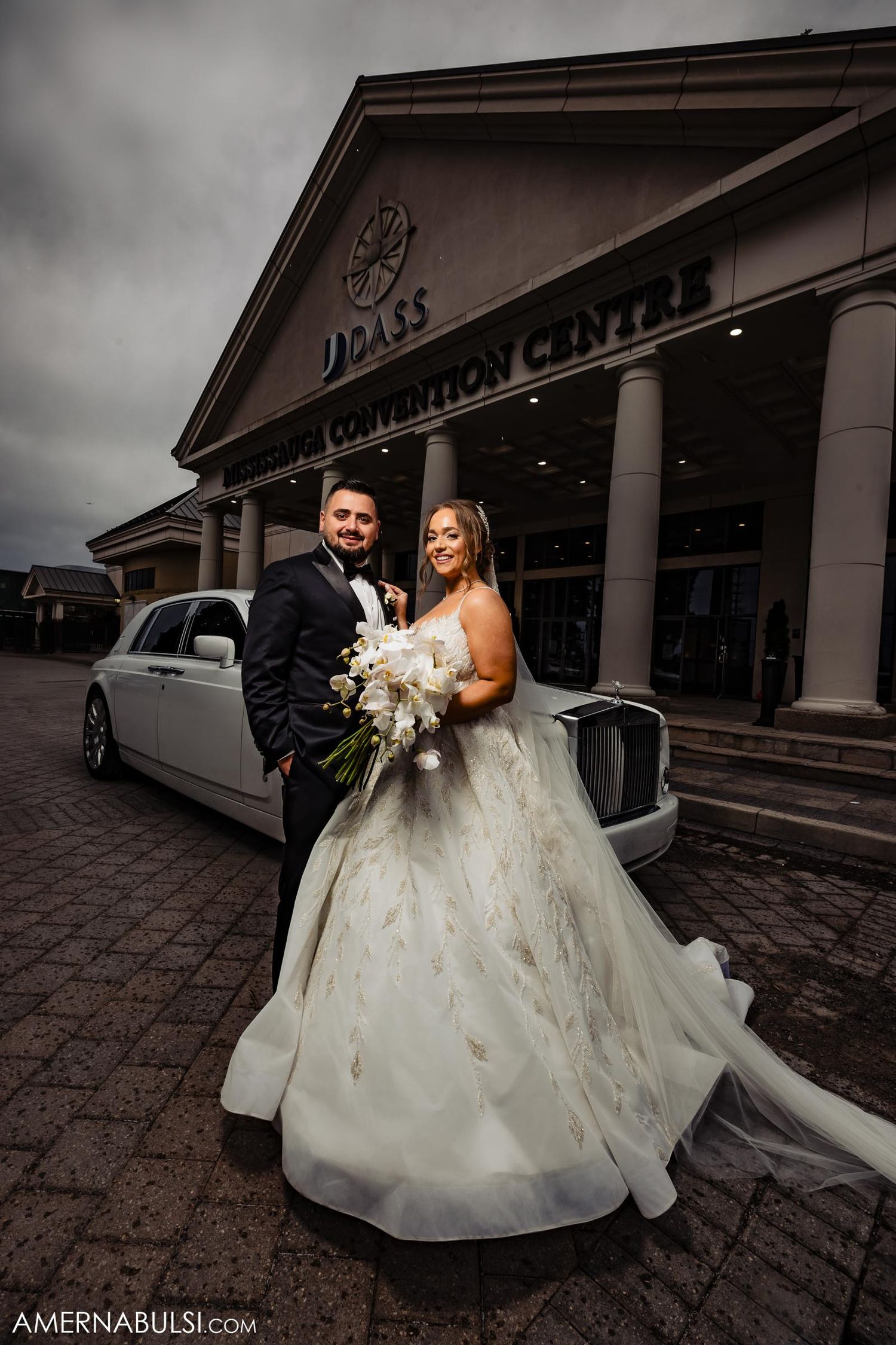 Wedding couple poses outside luxury venue with white limousine under dramatic cloudy sky.