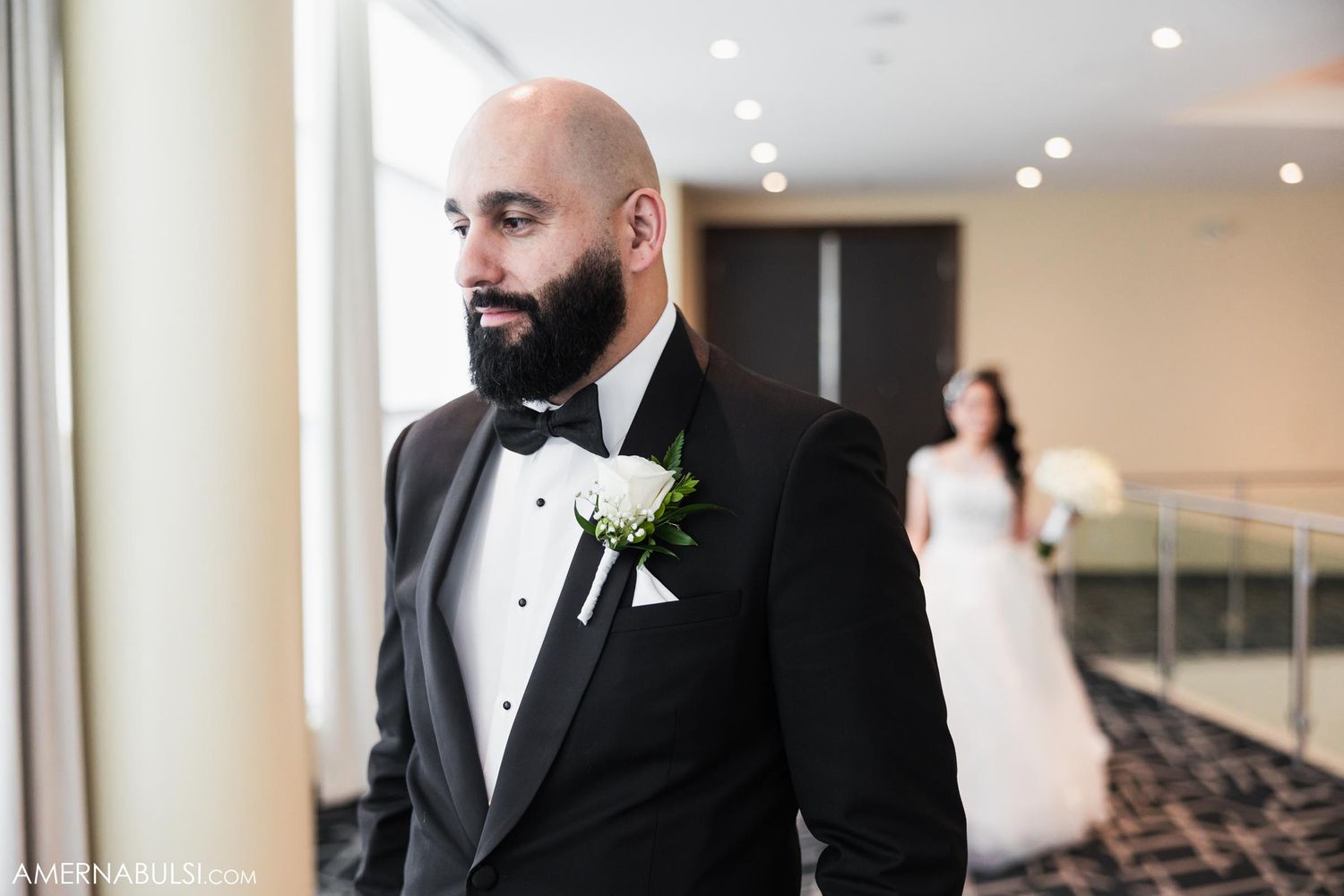 Groom in black tuxedo with white boutonniere stands by window as bride waits in background.