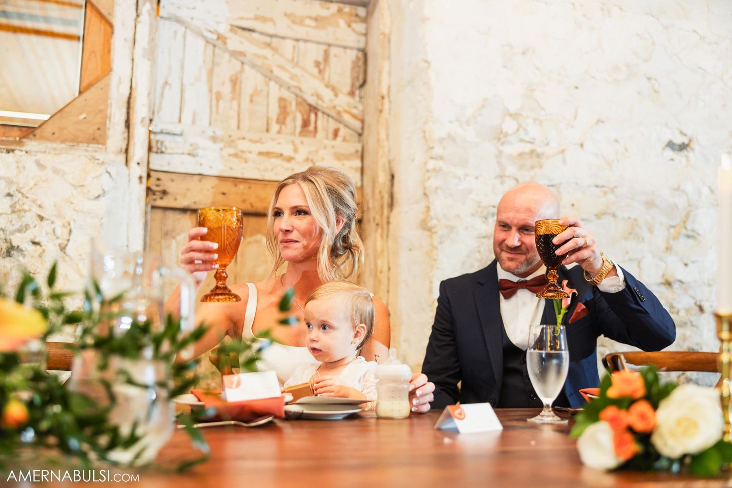 Wedding couple and child share meal at rustic reception while raising glasses in celebration.