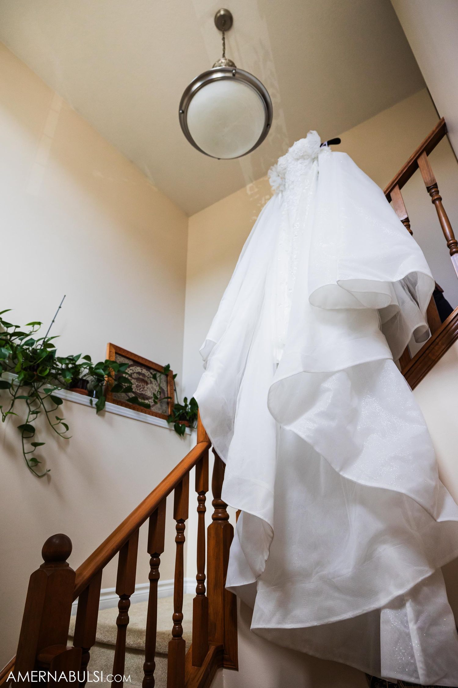 White wedding dress hanging in stairwell with circular light fixture and indoor plants.