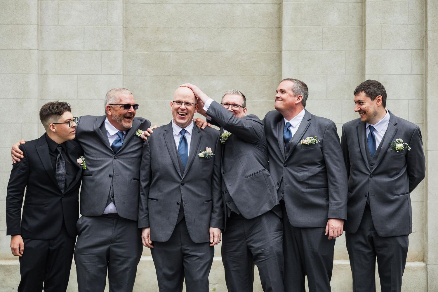 Groomsmen in dark suits pose together outdoors against light stone wall.