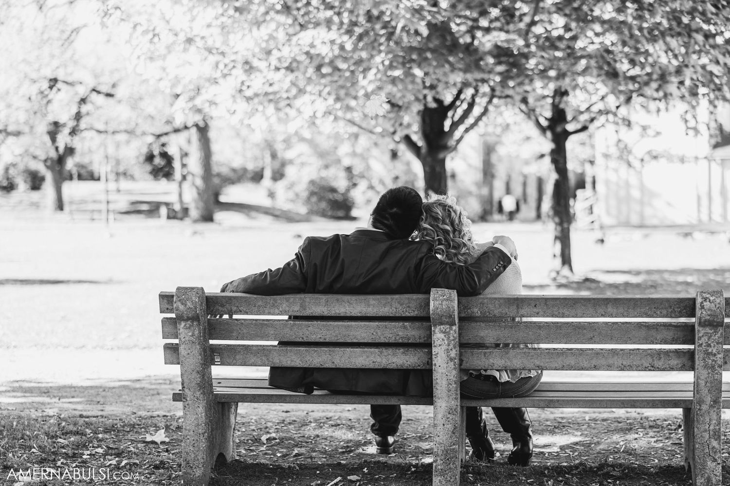 Black and white photo of couple sharing intimate moment on park bench under trees.