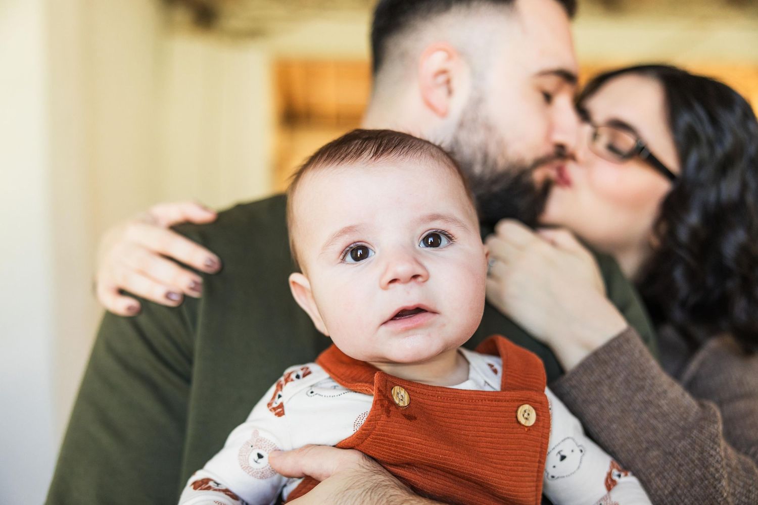 Intimate family moment with baby in orange romper while parents share tender moment.