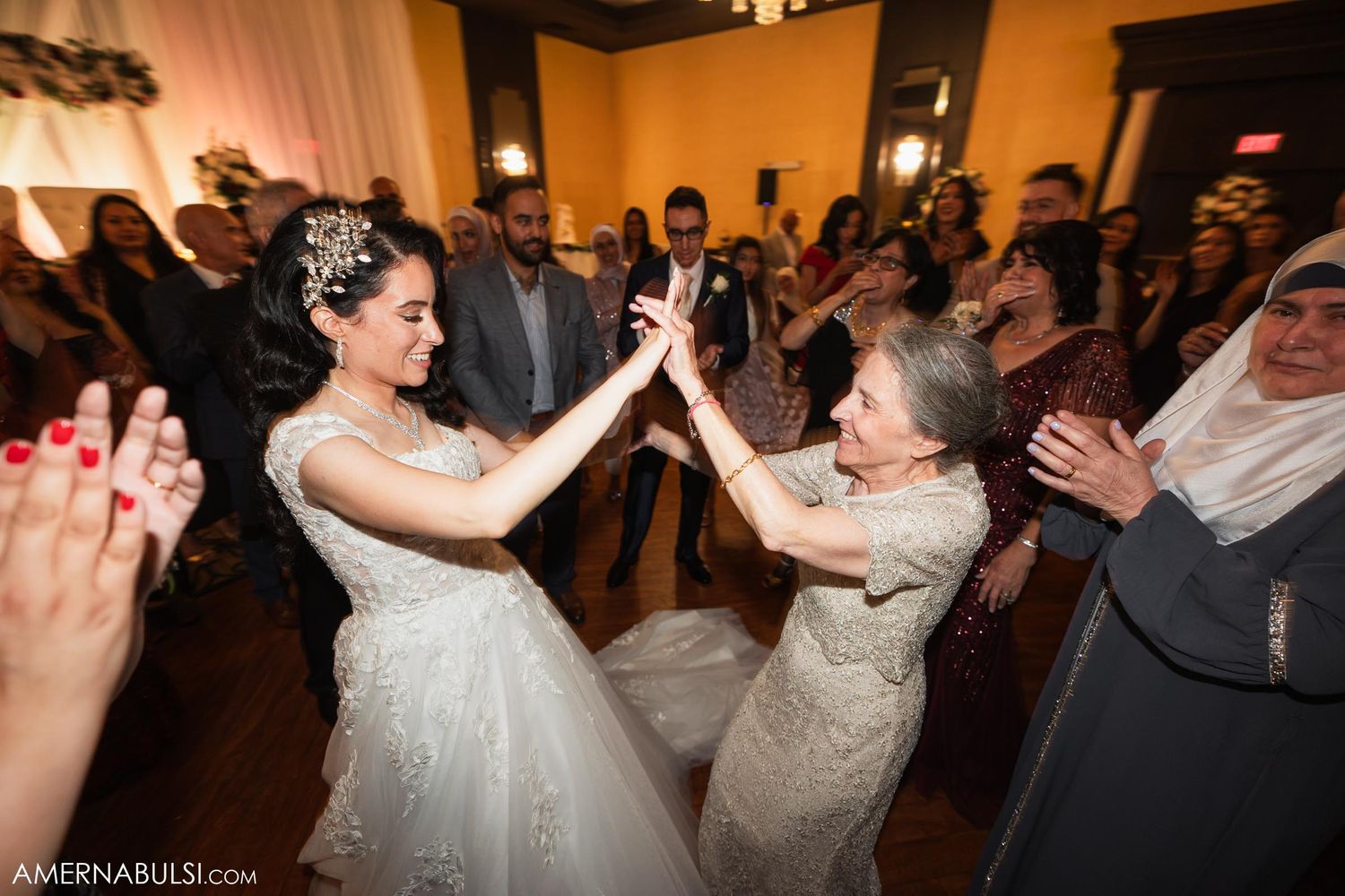 A bride shares a joyful dance with a guest while others clap and celebrate around them.
