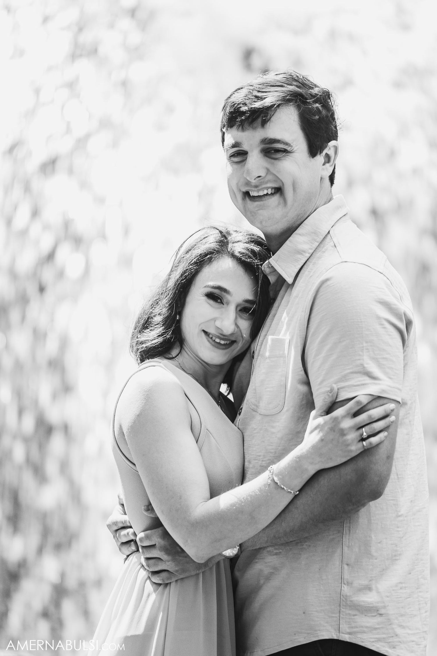 A black and white portrait of a couple embracing while smiling against a bright background.