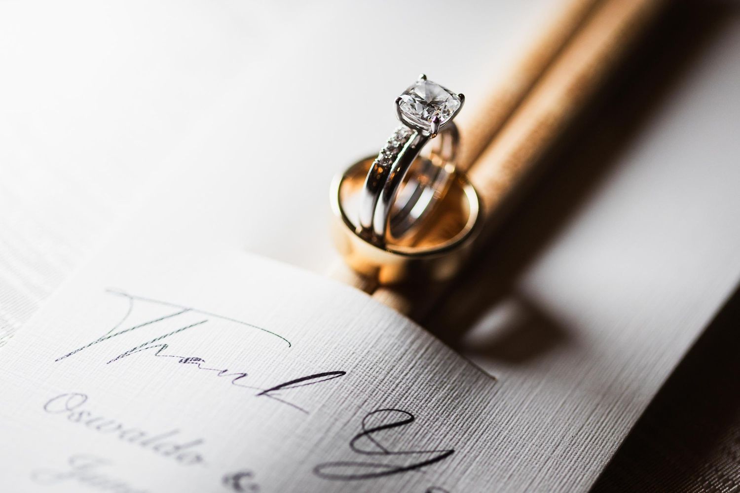 Wedding rings and diamond engagement ring placed on elegant invitation paper in dramatic lighting.