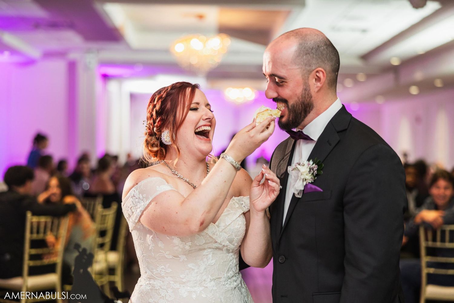 Bride feeding groom wedding cake while both laughing during reception.