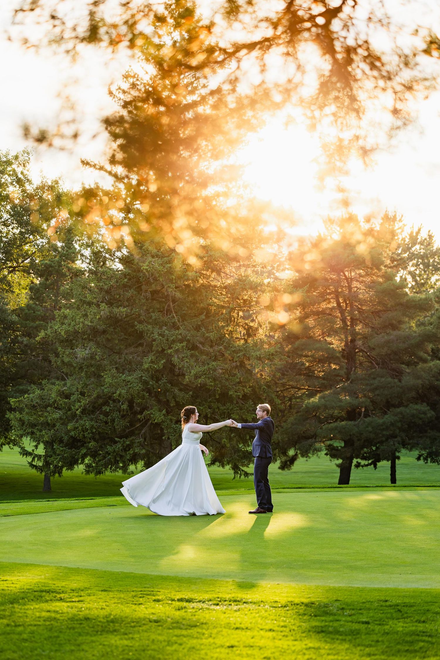 Couple twirls on manicured golf course as sunset filters through trees creating magical atmosphere.