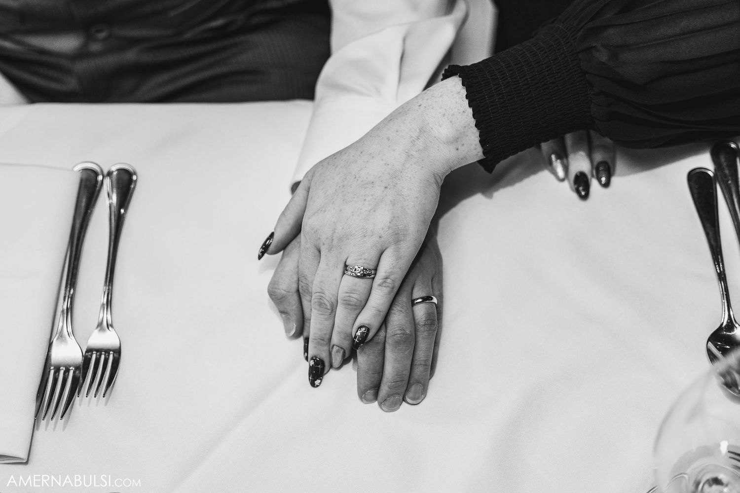 Close-up black and white photo of hands resting on a formal dining table setting with silverware.