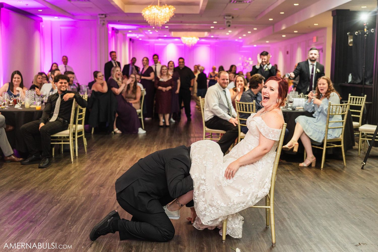 Traditional wedding garter removal scene during reception with guests watching.