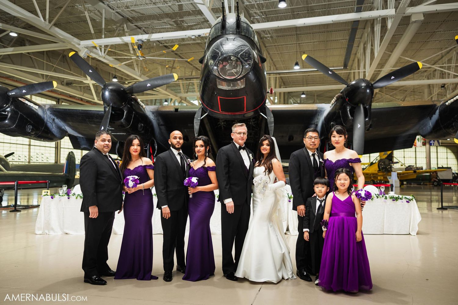 A wedding party poses in purple and black formal attire in front of a vintage military aircraft.