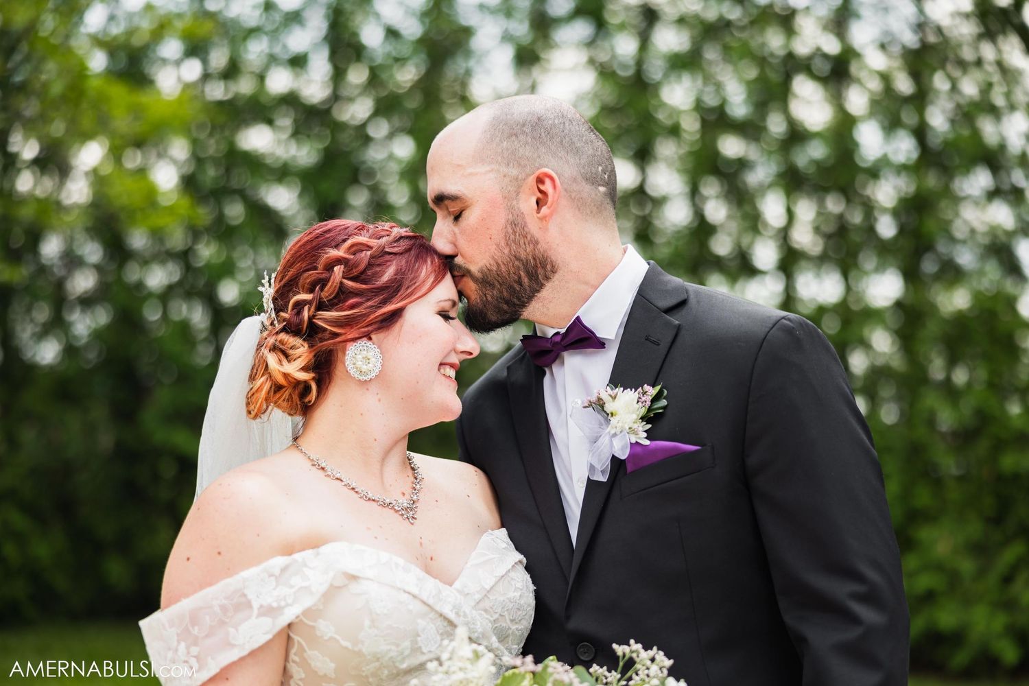 A romantic wedding portrait shows a tender moment between newlyweds against a natural green backdrop.