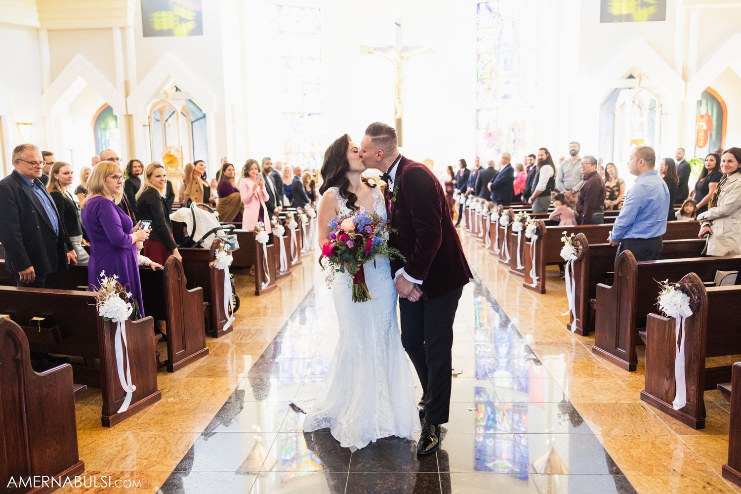 Romantic kiss in church aisle surrounded by wedding guests and natural light.