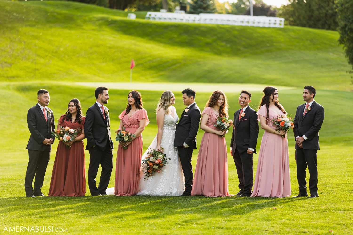 Bridal party in pink gowns and dark suits poses together on rolling golf course hills.