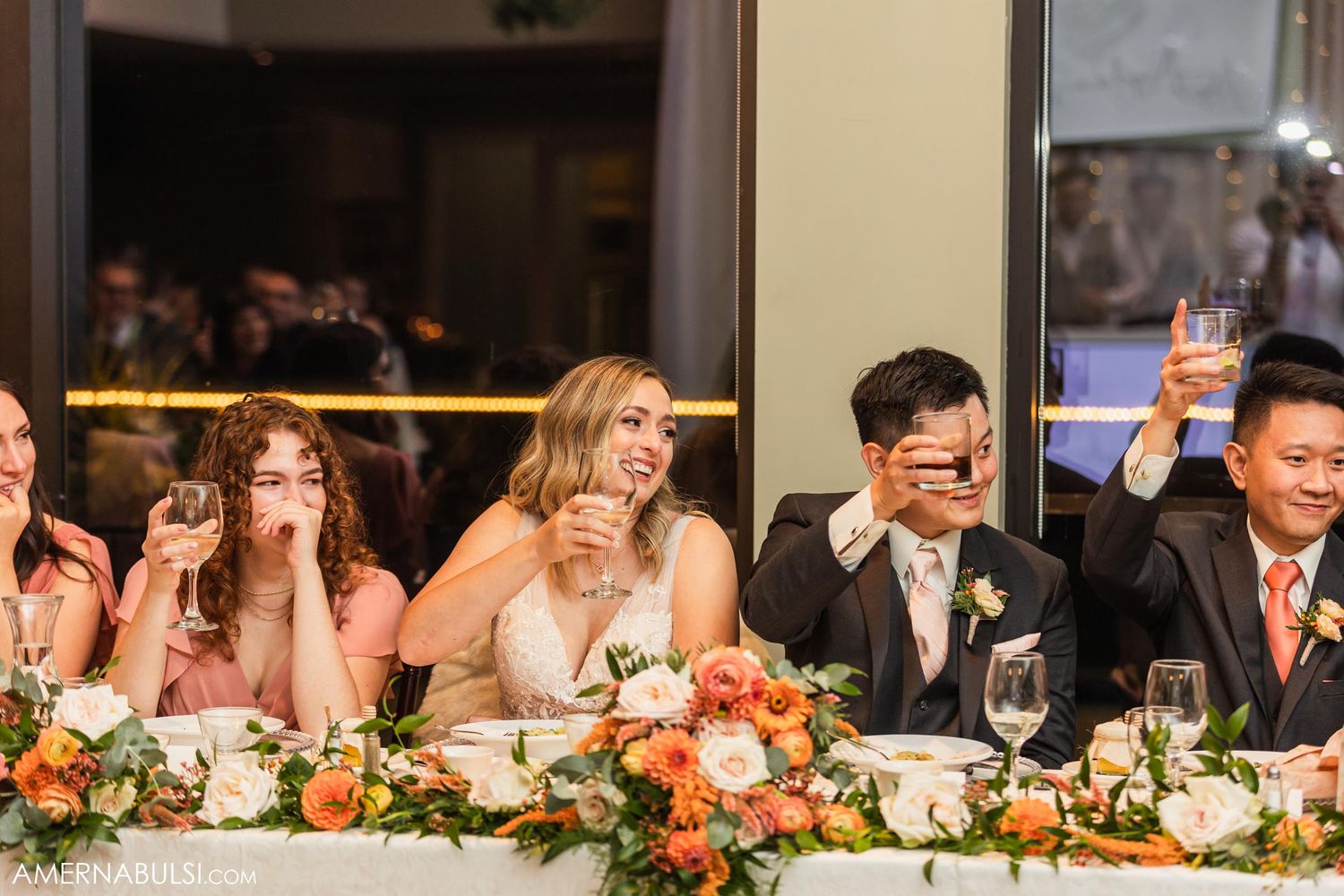 Wedding party raising glasses for a toast at decorated head table with floral arrangements.
