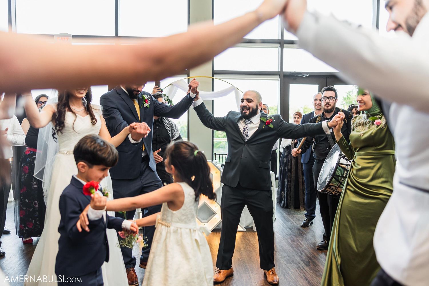 Guests form dance circle during wedding reception in bright venue with windows.