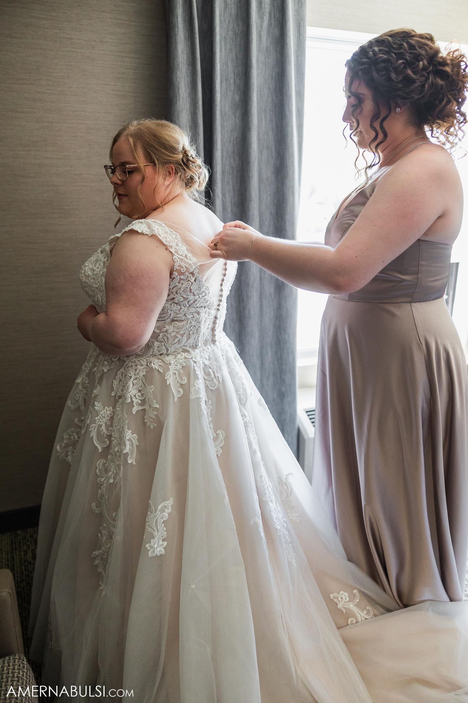 Helper assisting bride with the back of her detailed lace wedding gown.