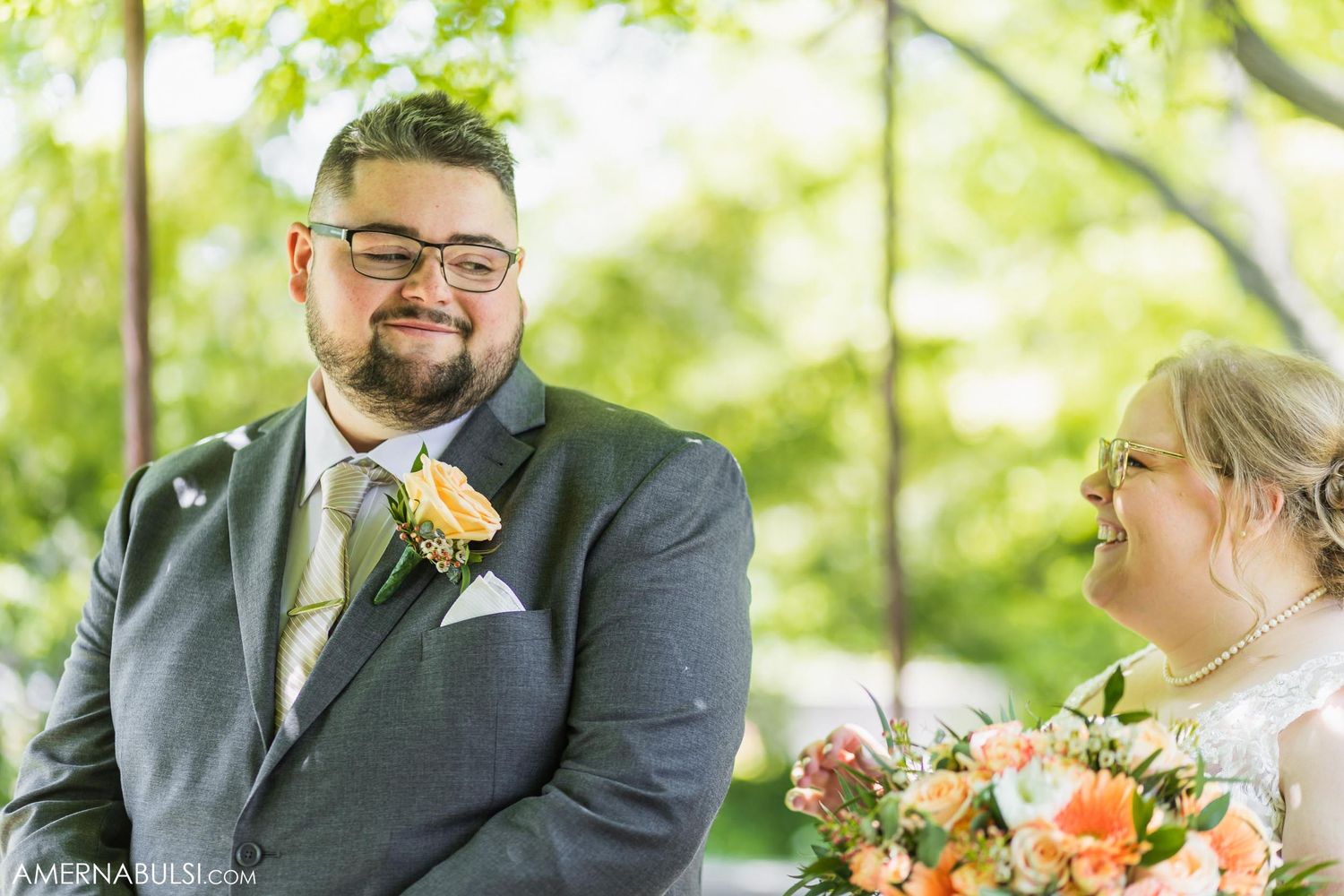 Wedding couple shares a joyful moment under green trees with peach-colored floral bouquet.