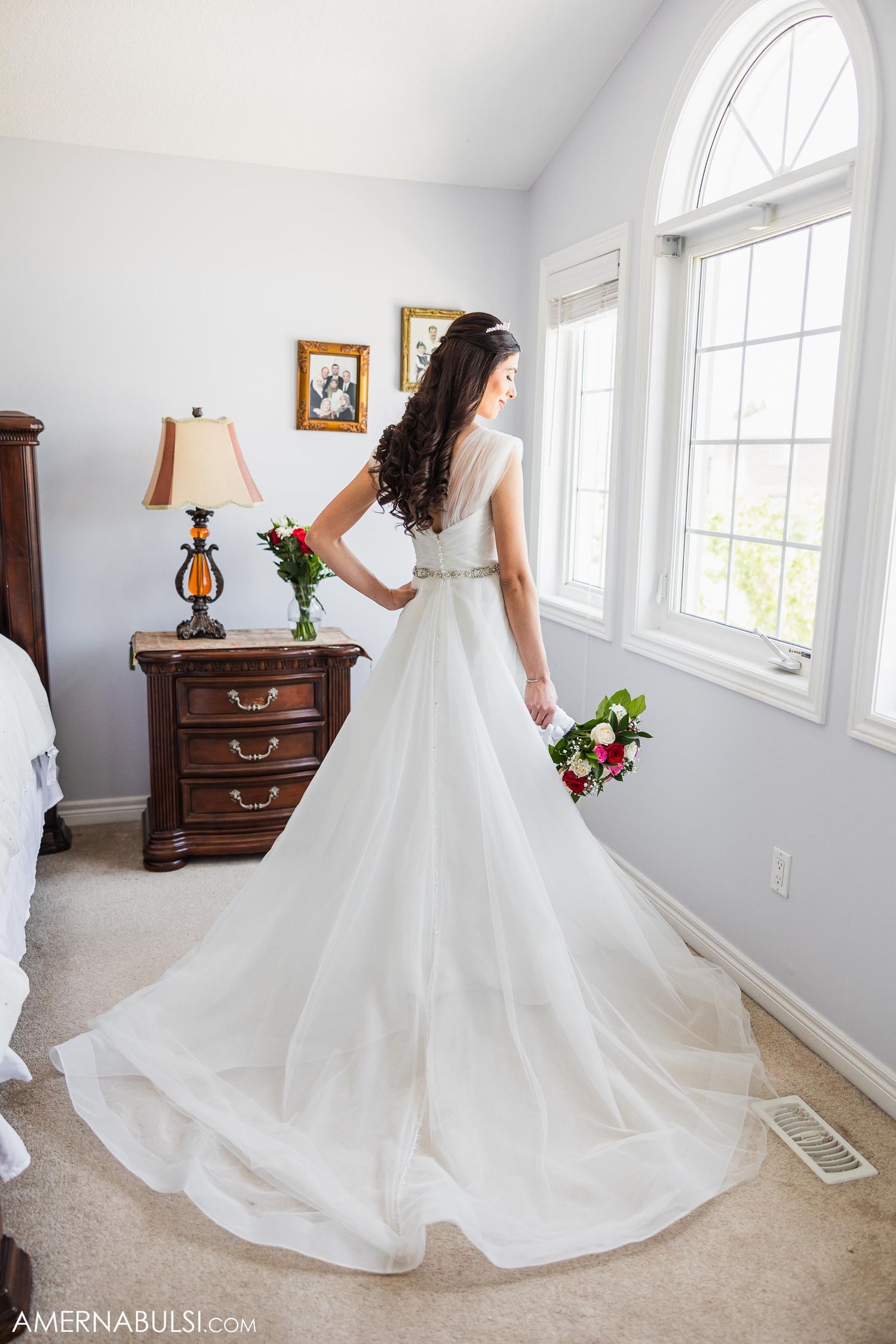 Elegant bride in flowing white gown poses by window while holding colorful bouquet.
