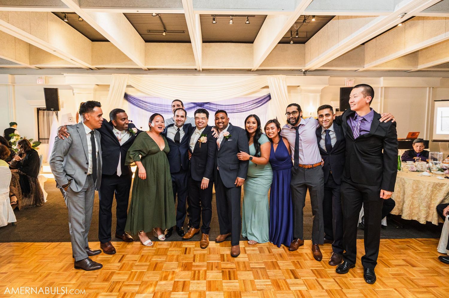 Wedding party poses for a group photo on parquet floor with decorative backdrop.