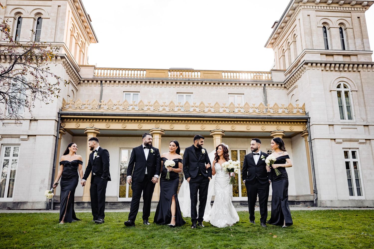 Wedding party poses in formal attire in front of a grand historic mansion.
