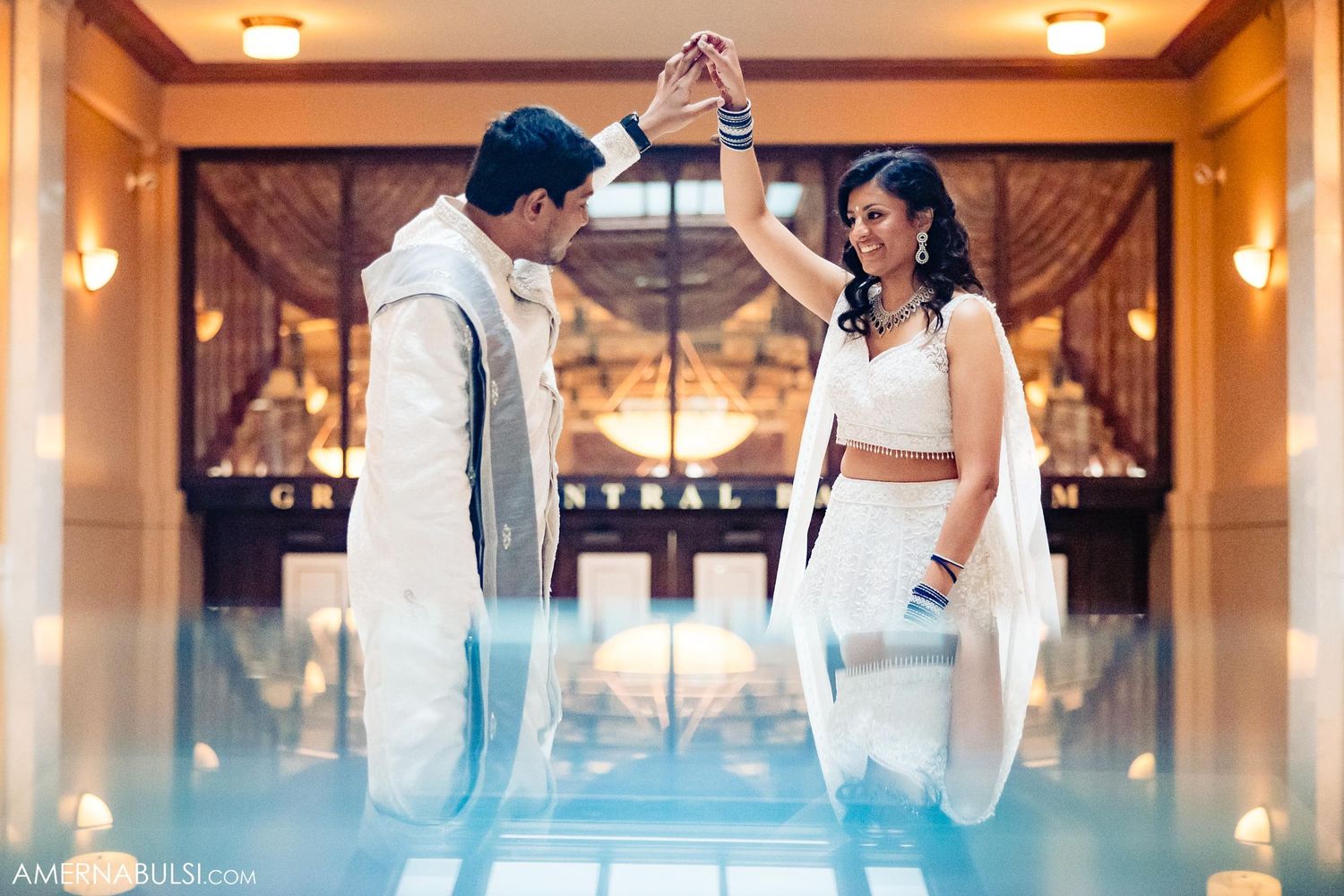 Couple in white wedding attire dance on reflective floor with warm lighting.