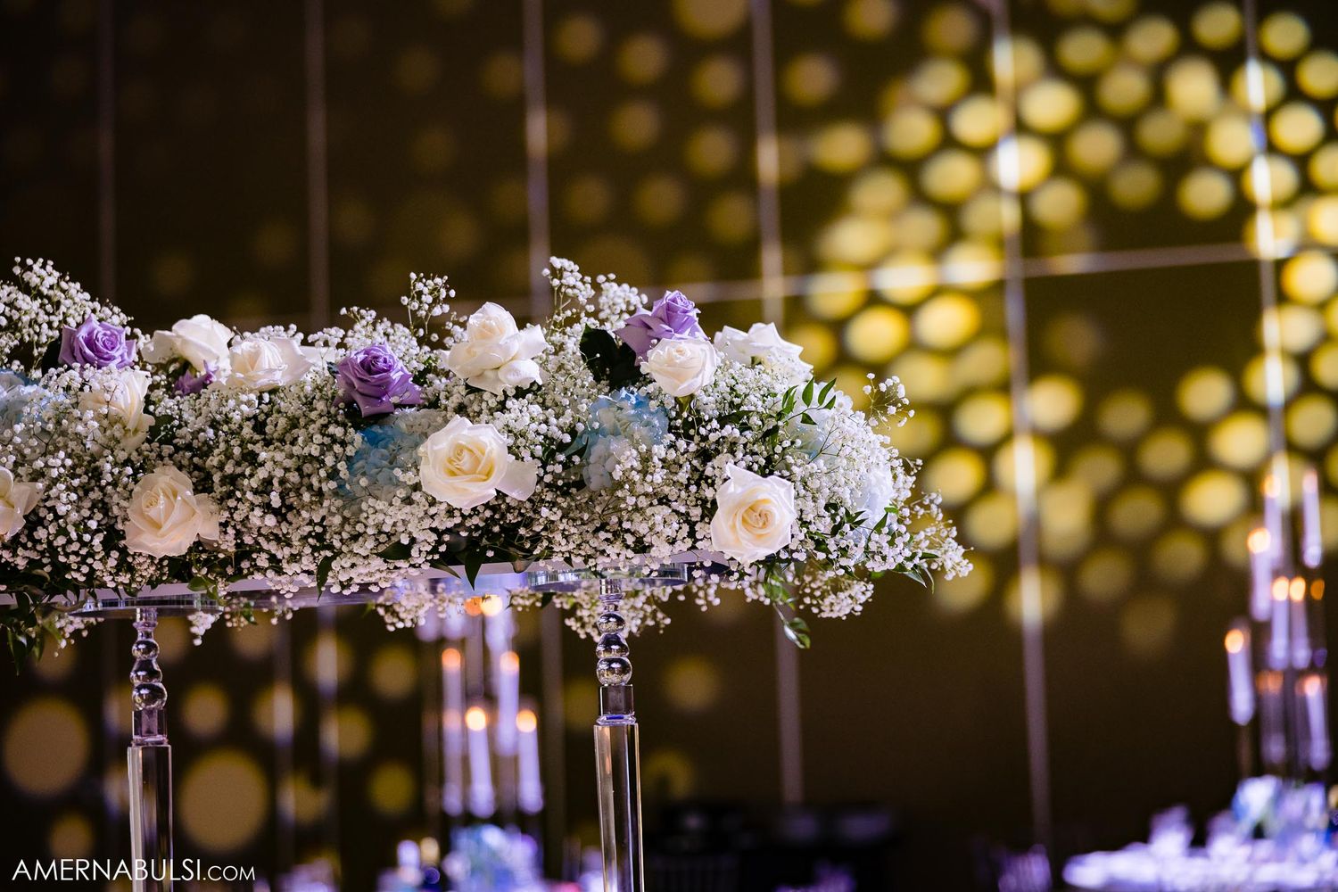 Elegant floral centerpiece with white and purple blooms against golden bokeh background.