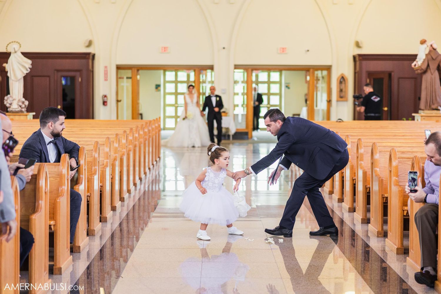Wedding attendant helps small flower girl in white dress walk down church aisle.