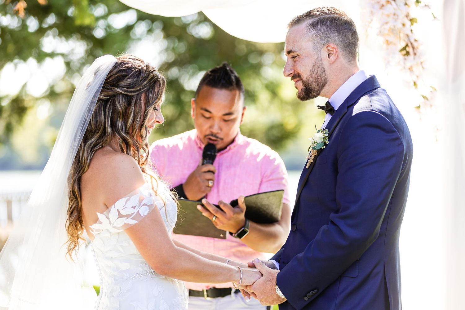 An outdoor wedding ceremony takes place under a wooden arch as the couple exchanges vows on a sunny day.