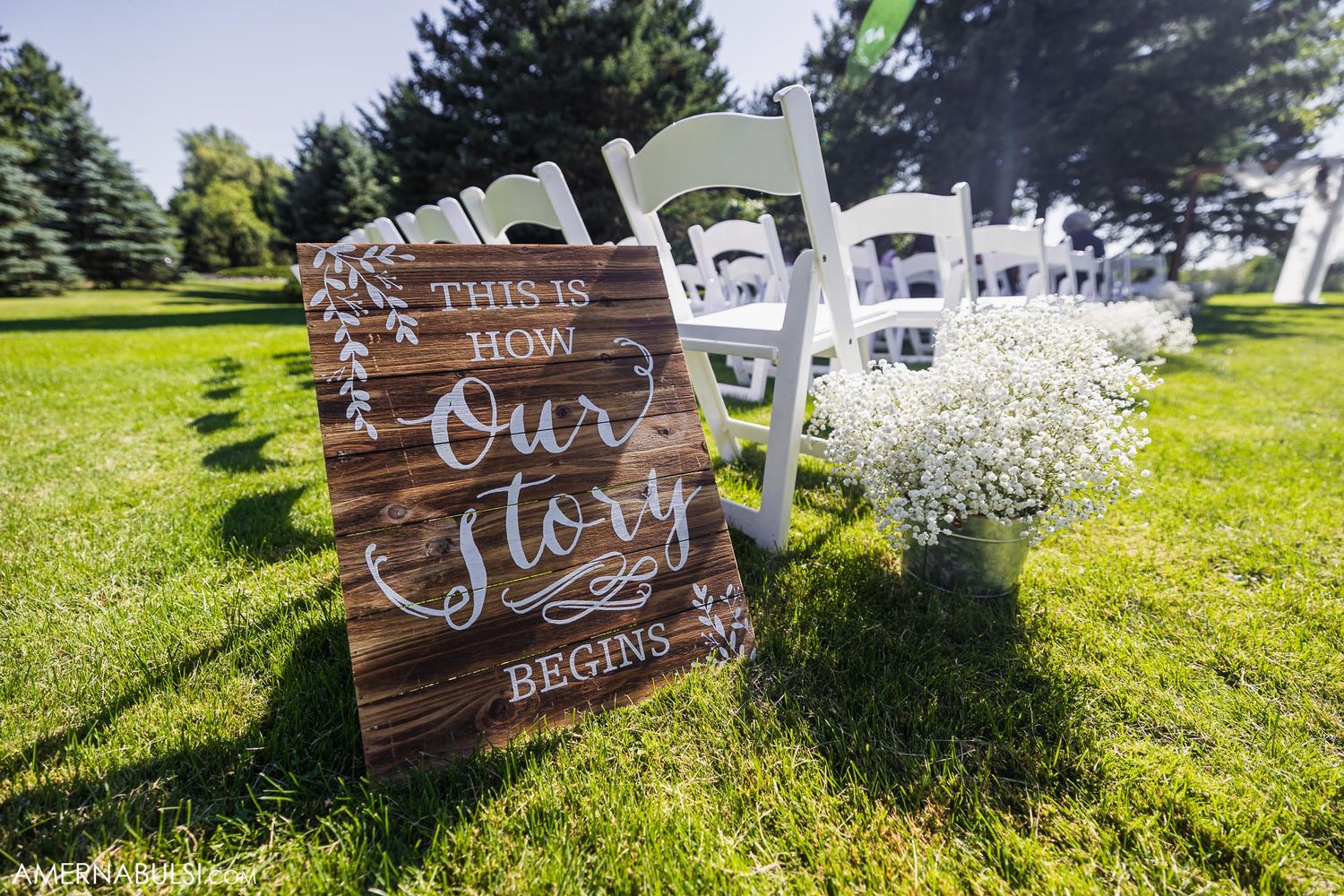 Rustic wooden wedding sign with white chairs and baby's breath flowers on grass.