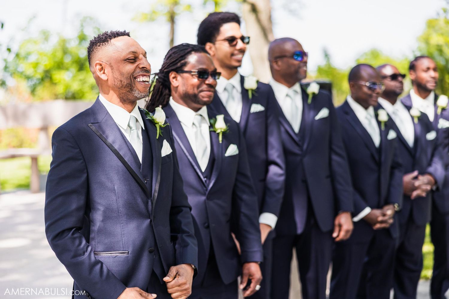 Line of groomsmen in matching navy suits laughing together outdoors.