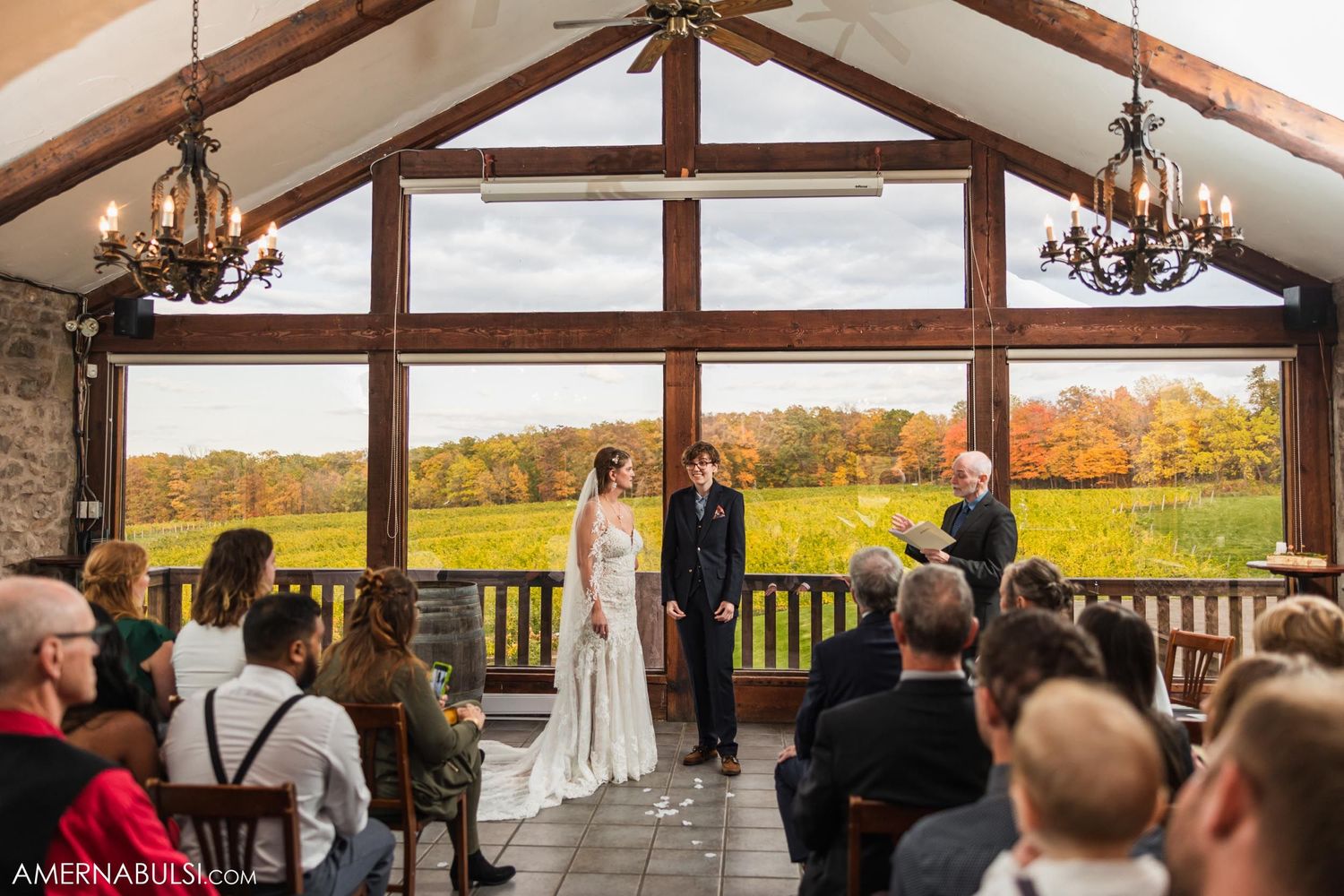 Guests gather in a rustic venue with vaulted ceilings and chandeliers for a wedding ceremony.
