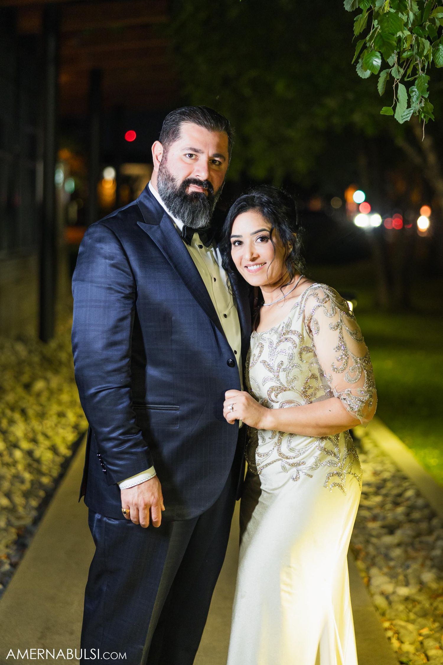 Elegantly dressed couple poses for romantic nighttime wedding portrait on illuminated pathway.